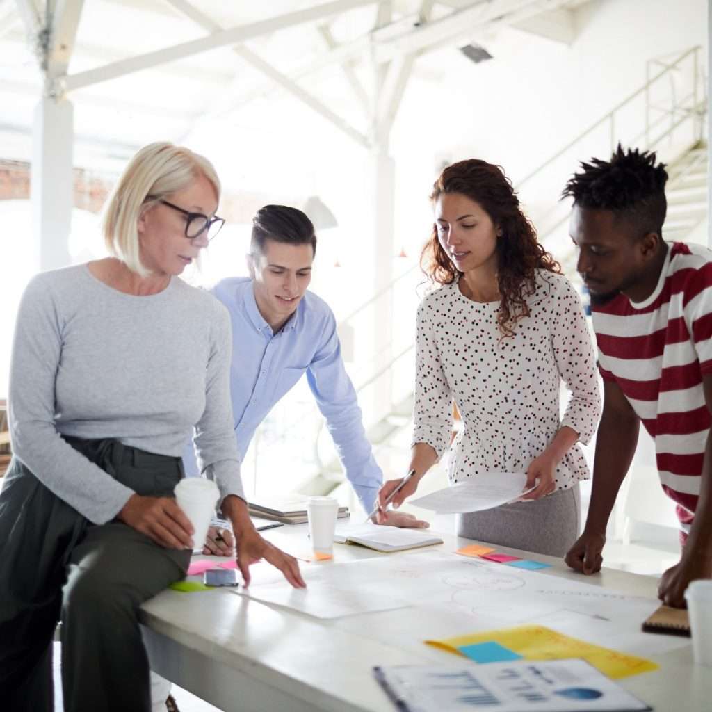A diverse group of people, including [mention the number] men and women of various ages and ethnicities, gather around a table in a professional setting. They appear engaged in a meeting, discussing plans and ideas with laptops, notebooks, and papers spread out before them.
