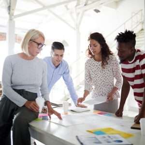 A diverse group of people, including [mention the number] men and women of various ages and ethnicities, gather around a table in a professional setting. They appear engaged in a meeting, discussing plans and ideas with laptops, notebooks, and papers spread out before them.
