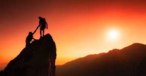 A strong leader, with a determined expression, pulls a person up a steep rock face. The person being pulled is reaching up to grasp the leader's outstretched hand.