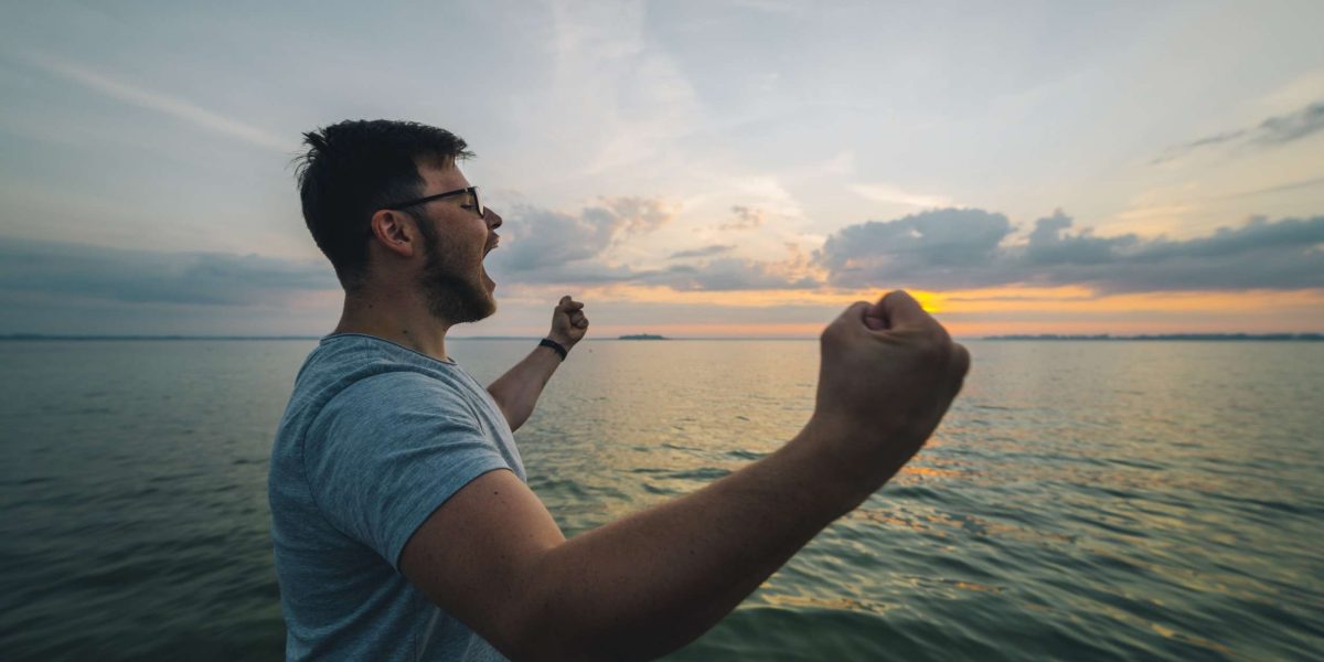 A silhouette of a person standing alone, looking out over a vast body of water with arms raised in a gesture of accomplishment. While the image captures a sense of individual achievement, the vastness of the water suggests humility in the grand scheme of things.